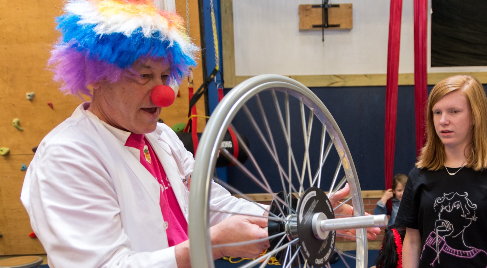 Evans Harrell in a rainbow clown wig, red nose, and lab coat, spinning a bicycle wheel to demonstrate angular momentum while a young woman watches