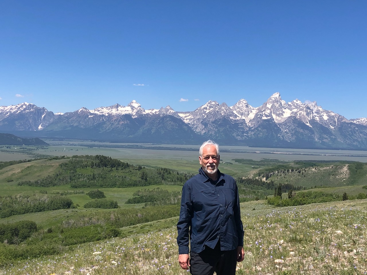 Evans Harrell standing in a wildflower meadow, the snow-capped Teton range and the Jackson Hole valley stretching out behind him